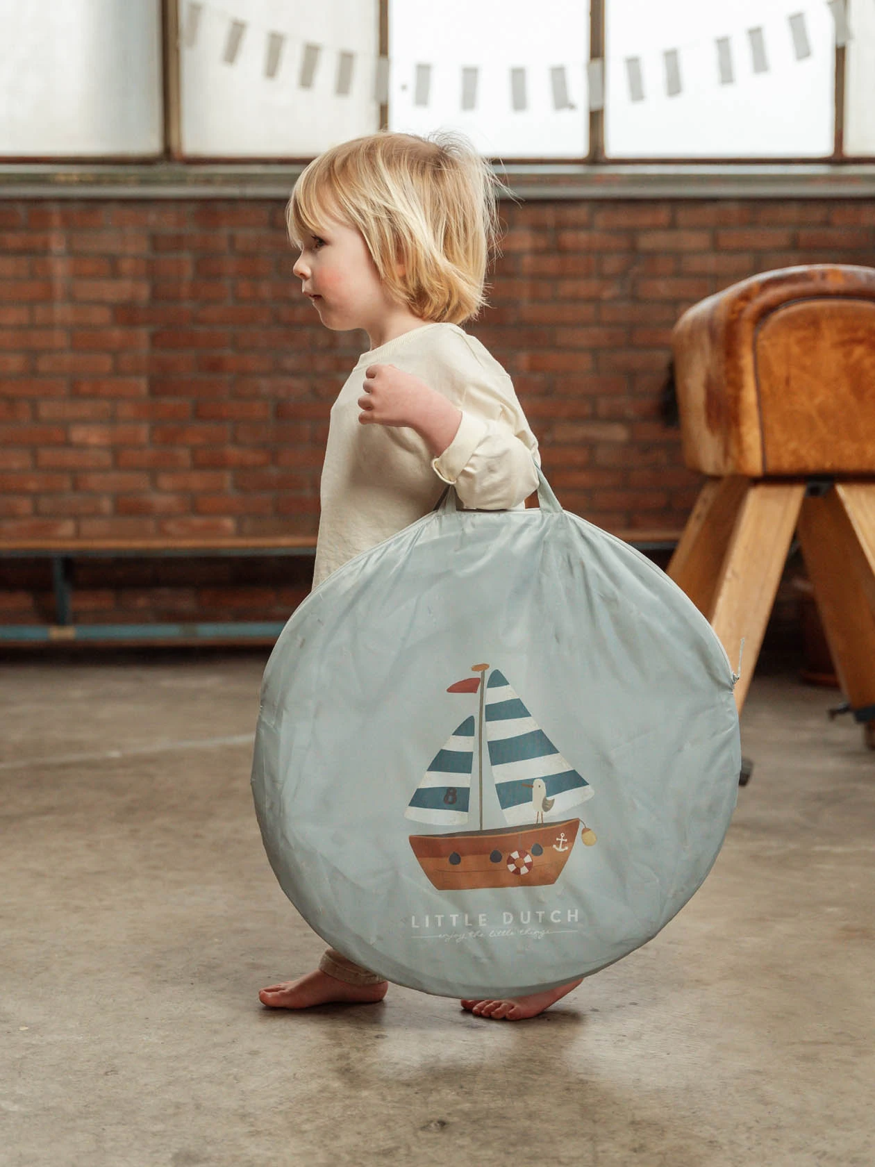 Małe dziecko niesie zapakowany w podręcznej torbie tunel dziecięcy z serii Sailors Bay.