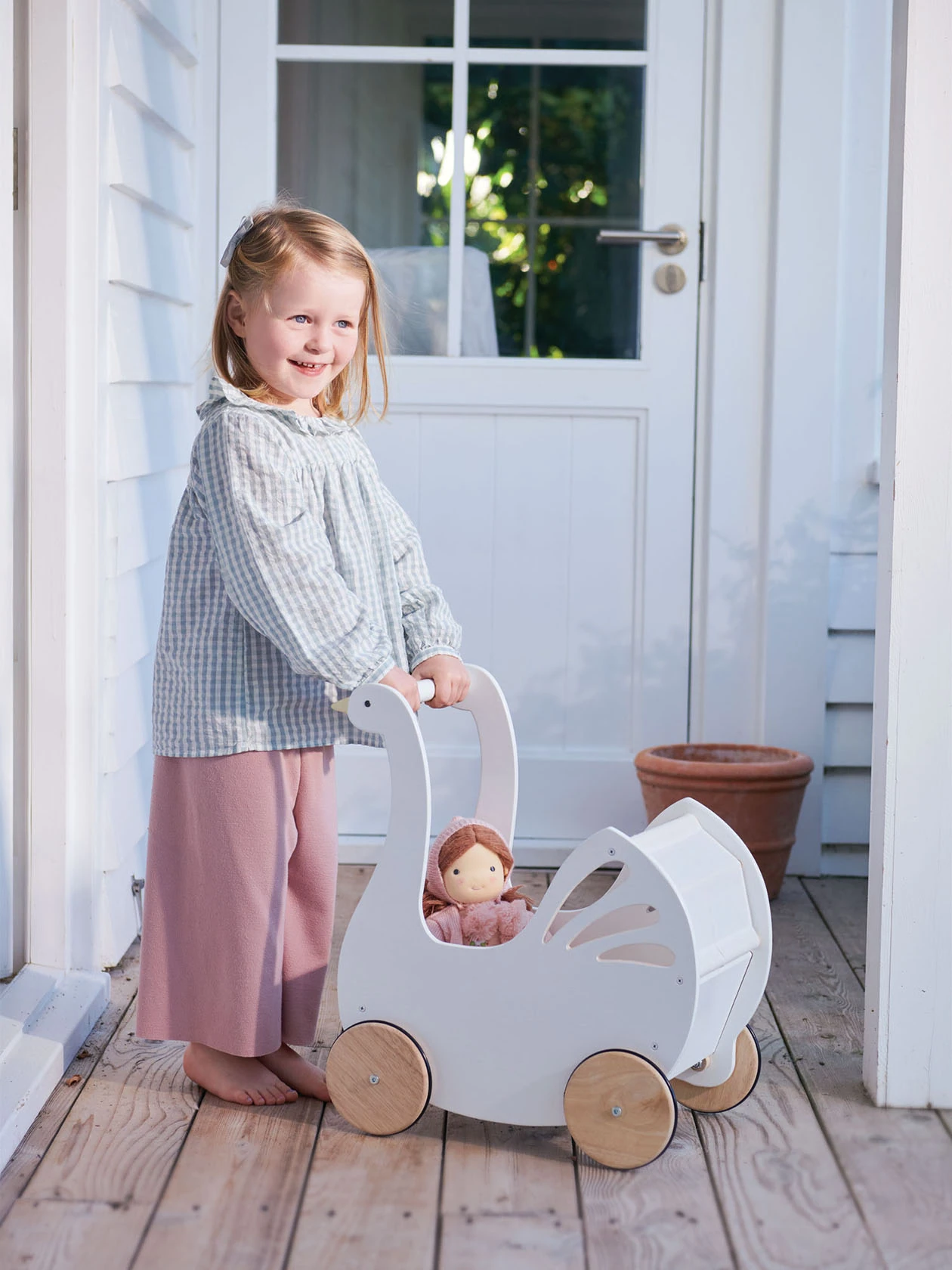 Mała dziewczynka stoi obok drewnianego wózka dla lalek Łabędź od Tender Leaf Toys.